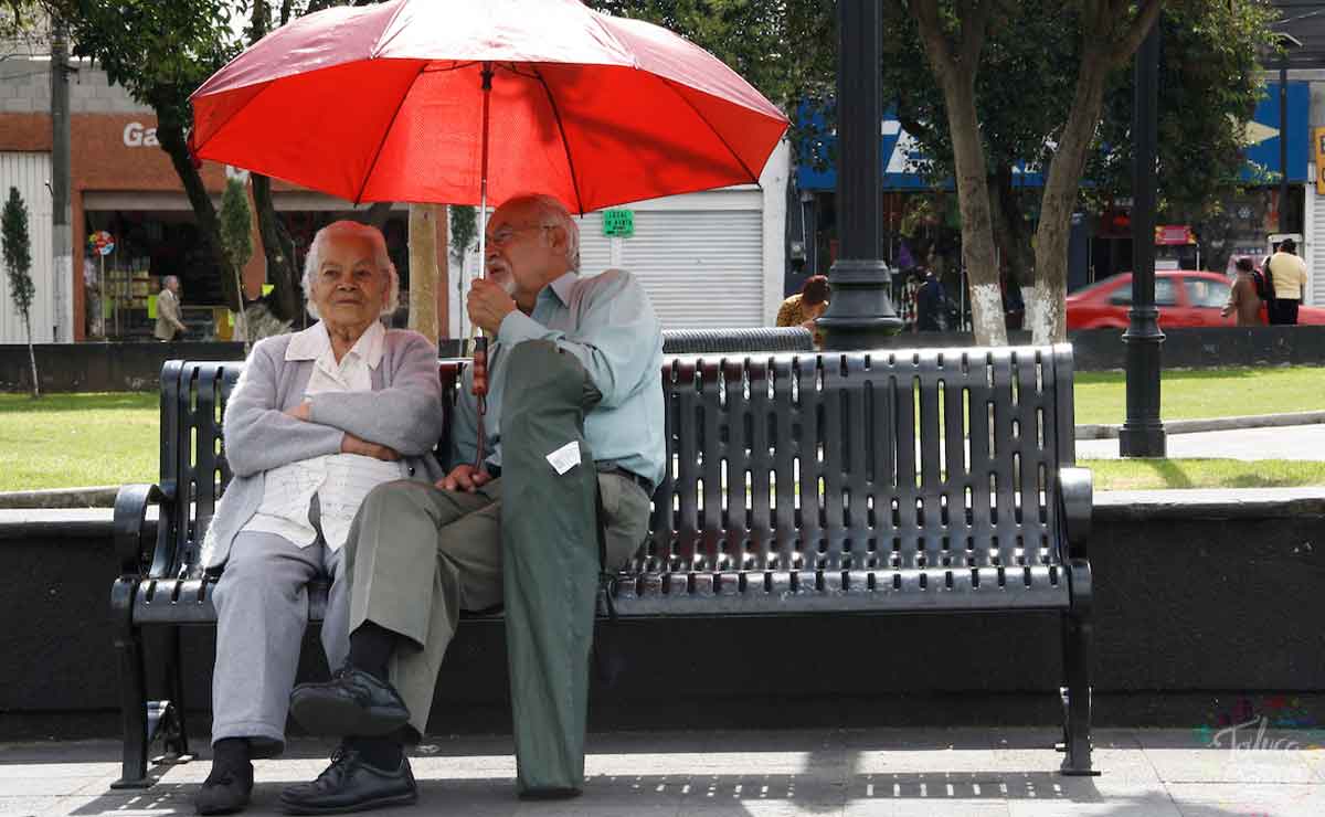 Dos adultos mayores que están sentados en una banca tapándose del sol y esperando su pensión bienestar