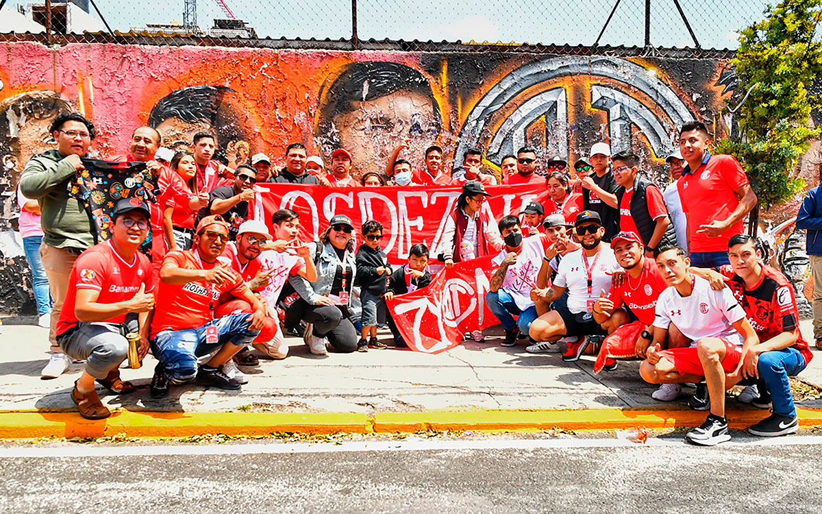 Grupo de personas aficionadas al futbol, posando cerca de un mural emblemático del club Toluca, ubicado en las afueras del estadio Nemesio Diez.